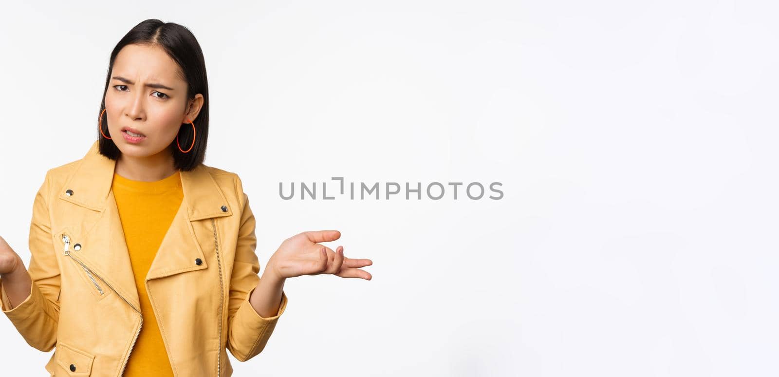 Image of confused young asian woman shrugging shoulders, looking puzzled and clueless at camera, cant understand, standing against white background.