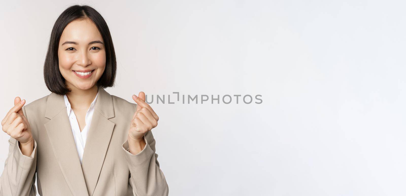 Cheerful asian saleswoman, smiling and showing finger hearts sign, standing in suit over white background.