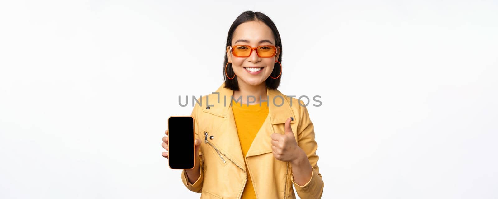 Beautiful korean girl, asian woman in sunglasses, showing smartphone app interface, thumbs up, recommending mobile phone application, white background.