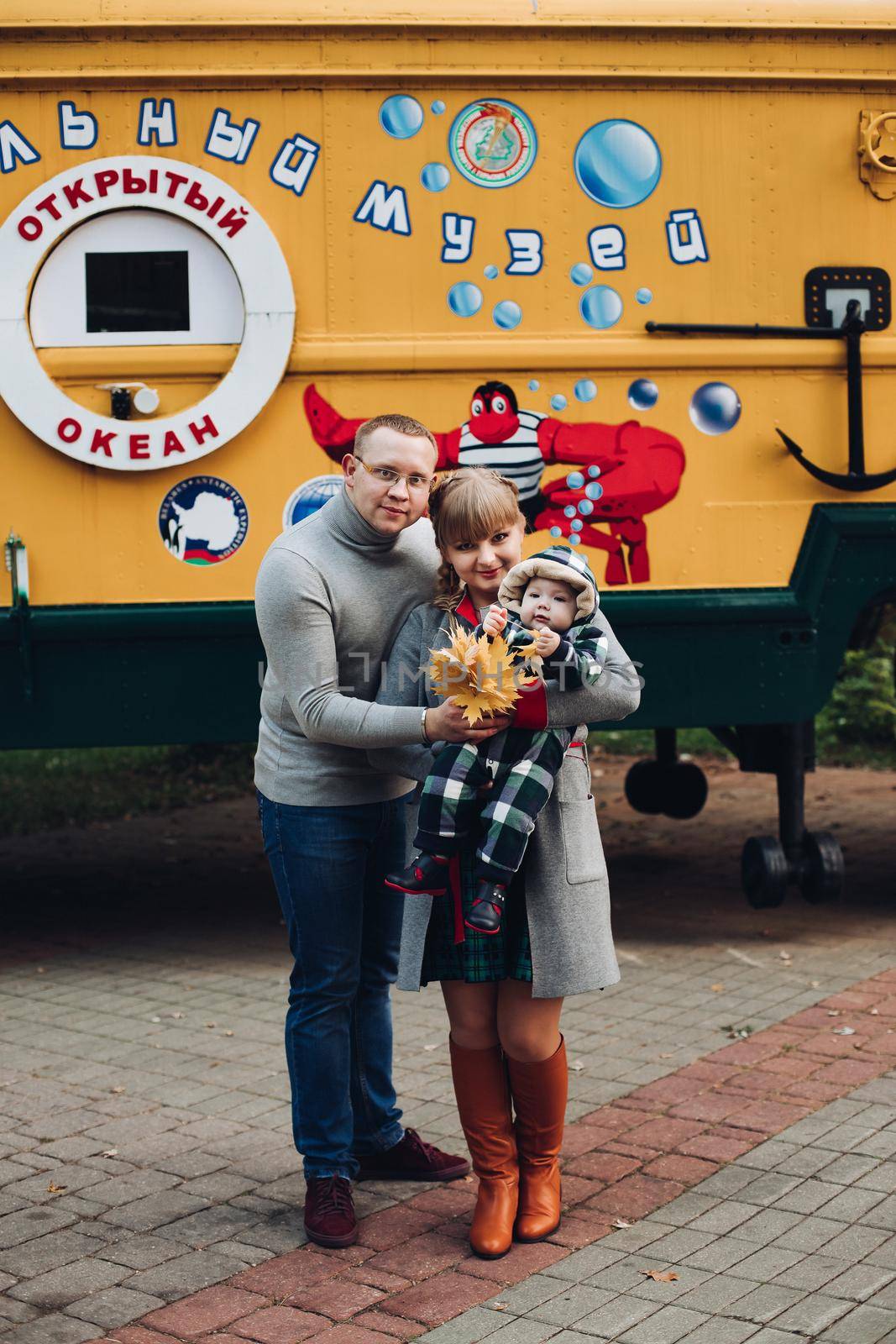 Portrait of attractive young mother and handsome smiling father wearing glasses holding their beautiful lovely baby girl on hands standing against green hedge in autumnal park. They are smiling and looking at camera.