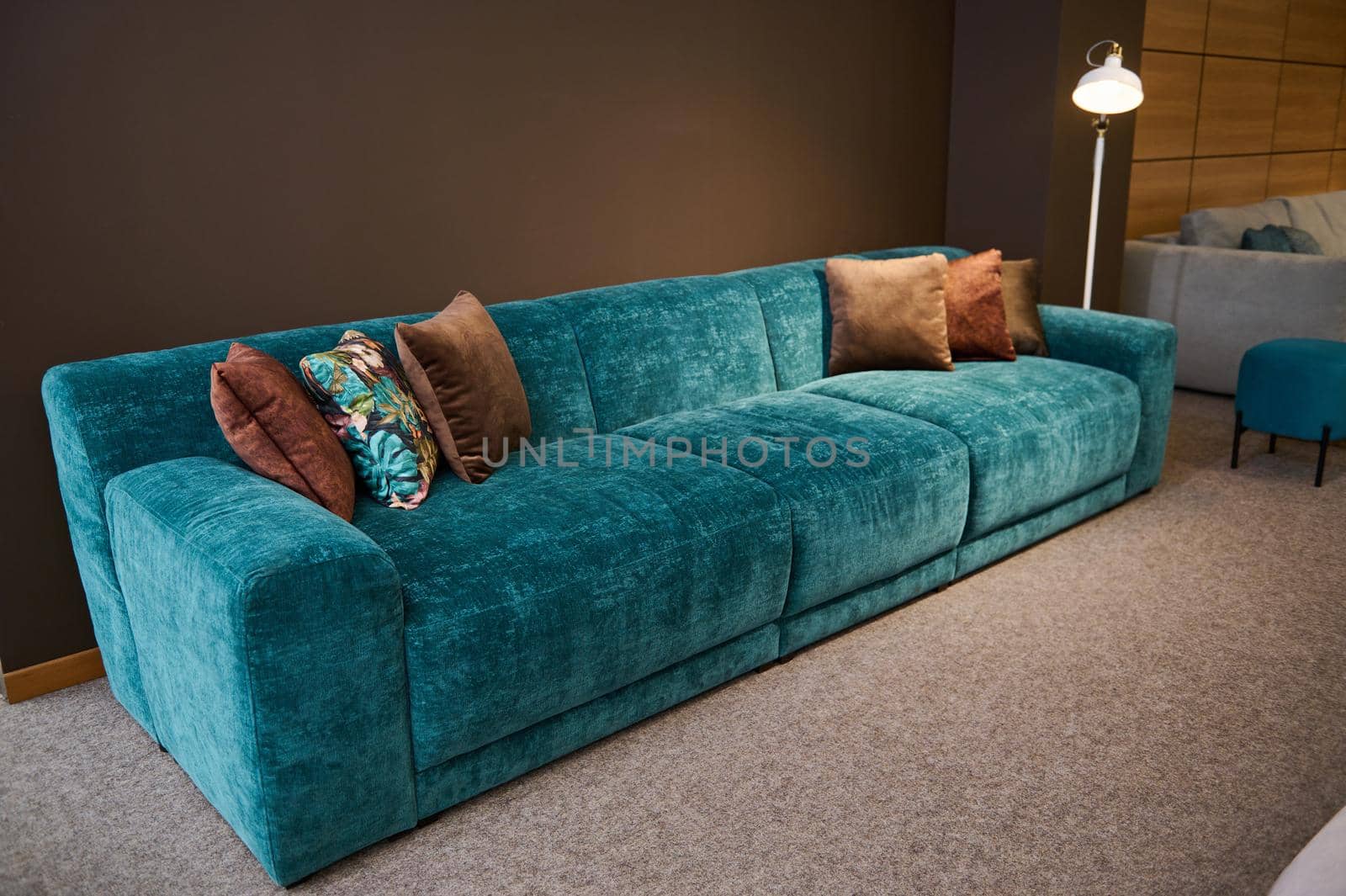 Exhibition of modern stylish upholstered furniture in the showroom of a furniture store. Focus on a turquoise soft velour sofa and brown cushions lit by a lamp against a brown wall background