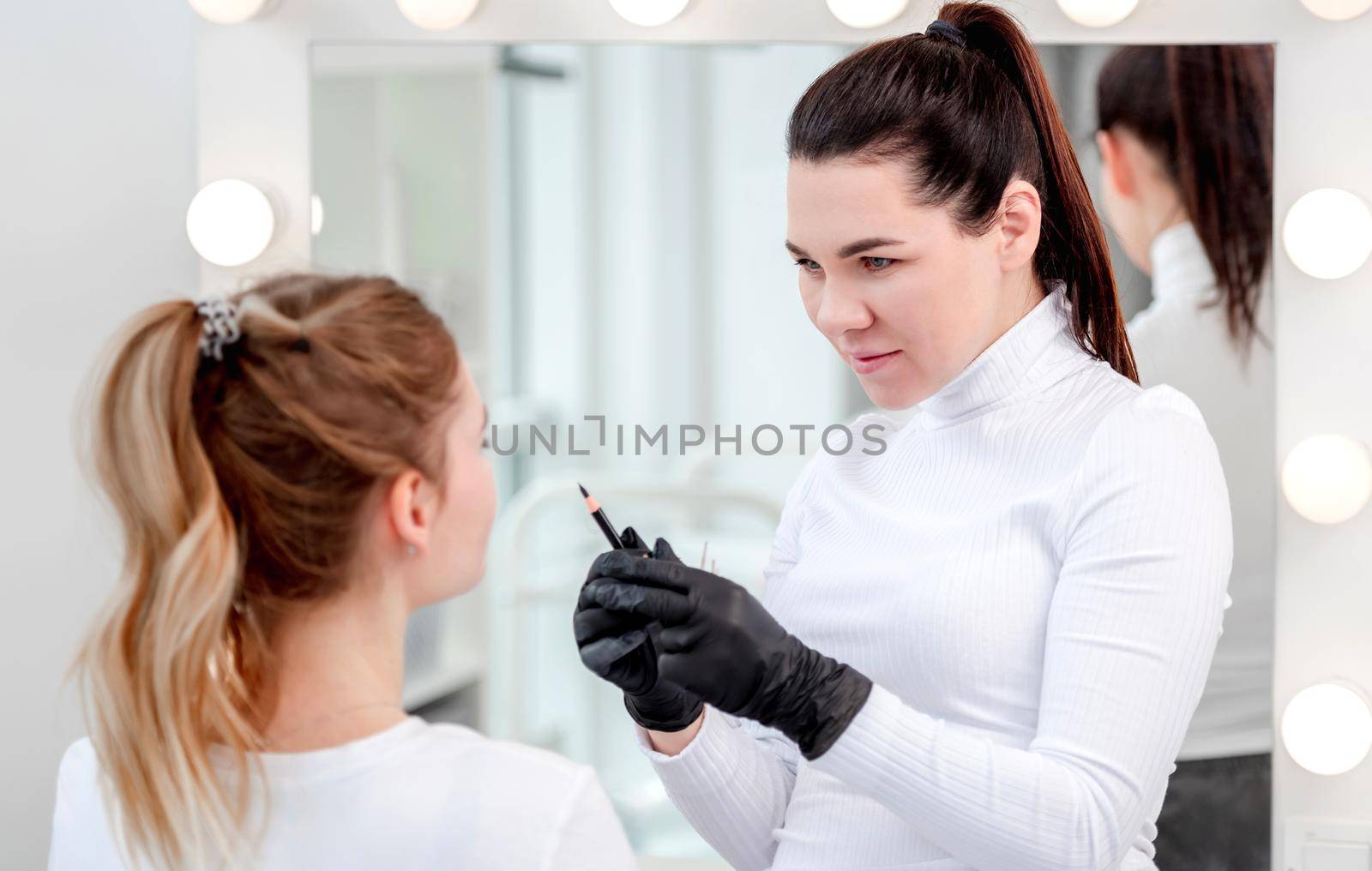 Permanent makeup master girl looking at model face during microblading process and holding professional tools in her hands