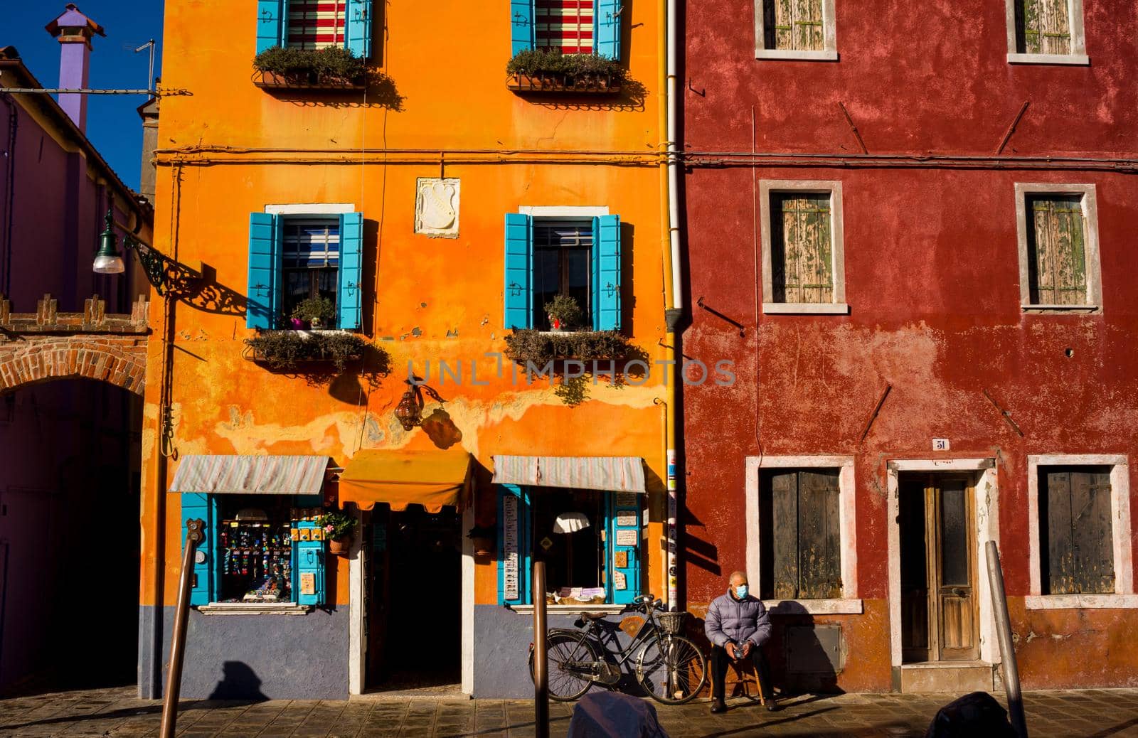 Italian elderly man sitting outside his house next his bicycle, behind the colorful houses of Burano island by bepsimage