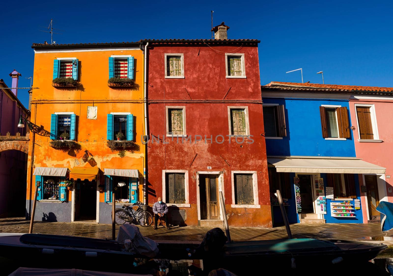 Burano, Italy- January, 06: Italian elderly man sitting outside his house next his bicycle, behind the colorful houses of Burano island on January 06, 2022