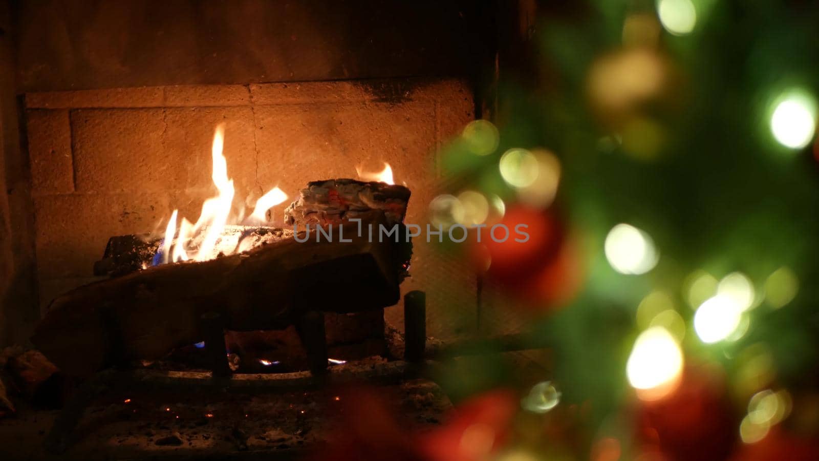 Christmas tree lights by fire in fireplace, New Years Eve or Xmas decoration of pine or fir with red balls. Festive fireside on winter christmastime holiday. Cozy place by burning firewood in december