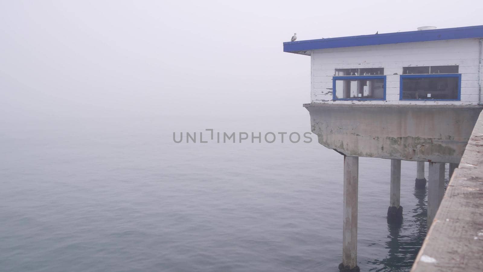 Wooden house on piles, water surface in haze, Ocean Beach pier, foggy California coast, USA. Old abandoned obsolete boardwalk cafe in misty weather. Calm tranquil silent atmosphere on San Diego shore.