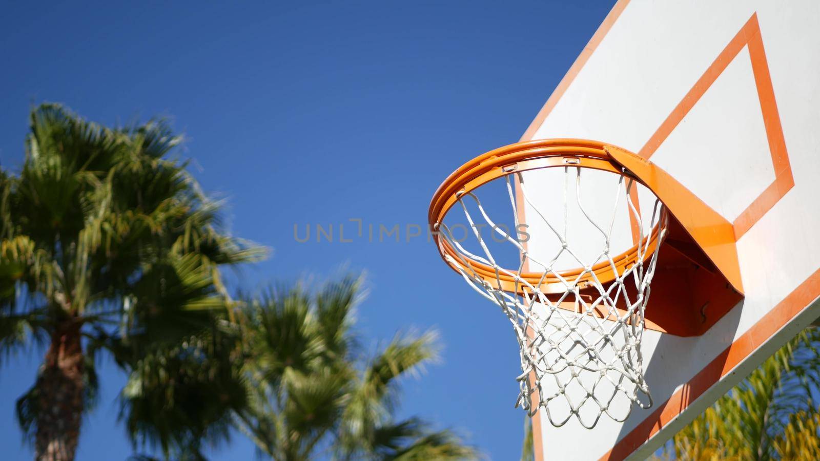 Basketball court outdoors, orange hoop, net and backboard for basket ball game outside. Recreational sport equipment on streetball field or playground. Blue sky and beach palm trees, California USA.
