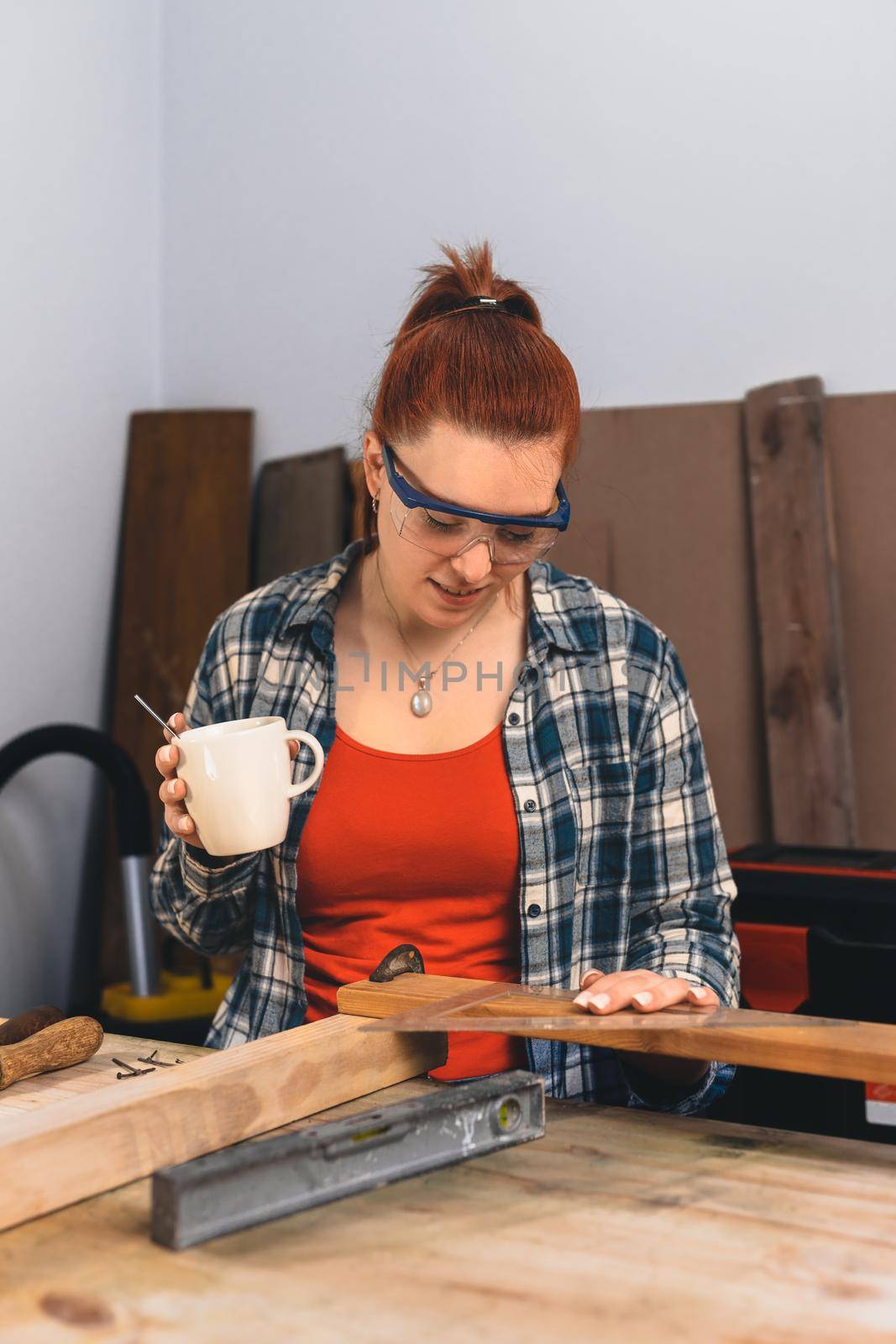 Young red-haired female carpenter having a coffee, planning her new project in her small carpentry business. 4 by CatPhotography