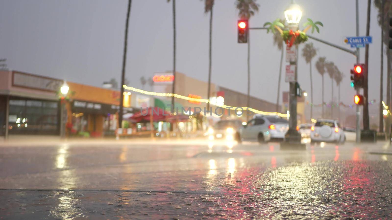 Cars lights reflection on road in rainy weather. Rain drops on wet asphalt of city street in USA, water raindrops falling on sidewalk. Palm trees and rainfall, twilight dusk. Ocean Beach, California.