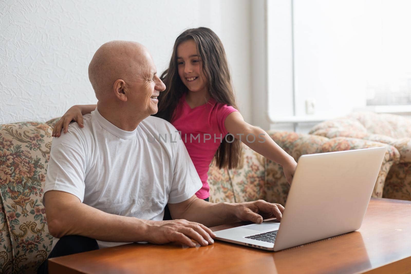 Happy little girl hugging smiling grandfather sitting on sofa with laptop