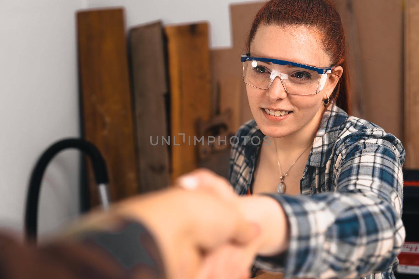 Young red-haired female carpenter shaking hands with a customer to close a deal. by CatPhotography