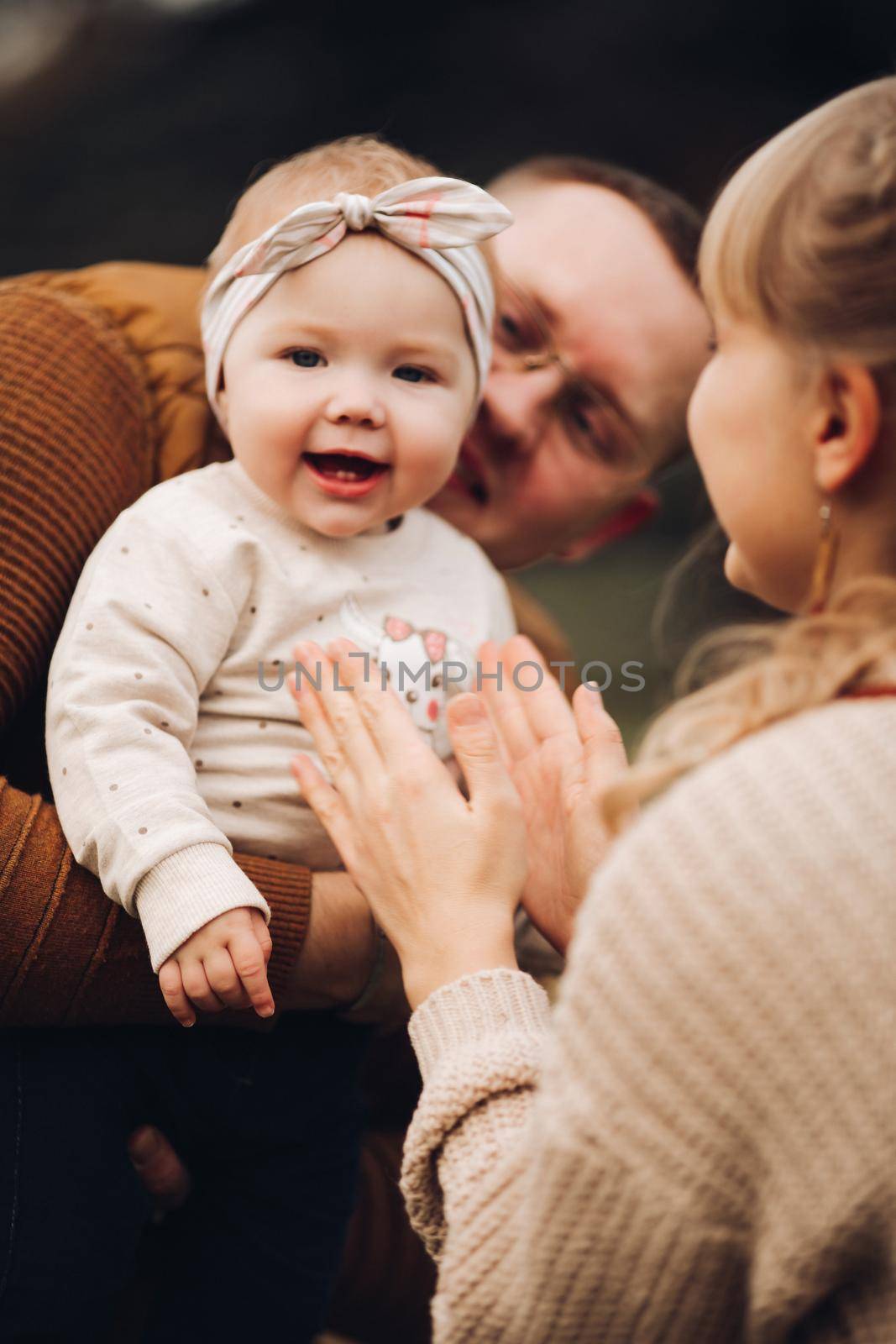 Portrait of attractive young mother and handsome smiling father wearing glasses holding their beautiful lovely baby girl on hands standing against green hedge in autumnal park. They are smiling and looking at camera.