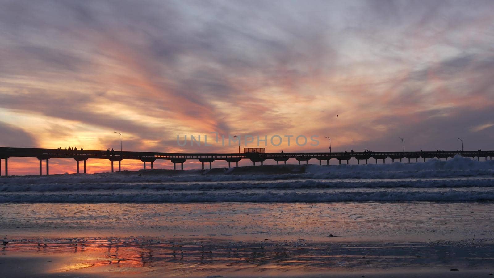 Silhouette of people walking, surfers surfing by pier in sea water. Ocean waves, dramatic sky at sunset. California coast, beach or shore vibes at sundown. Summer seascape seamless looped cinemagraph.