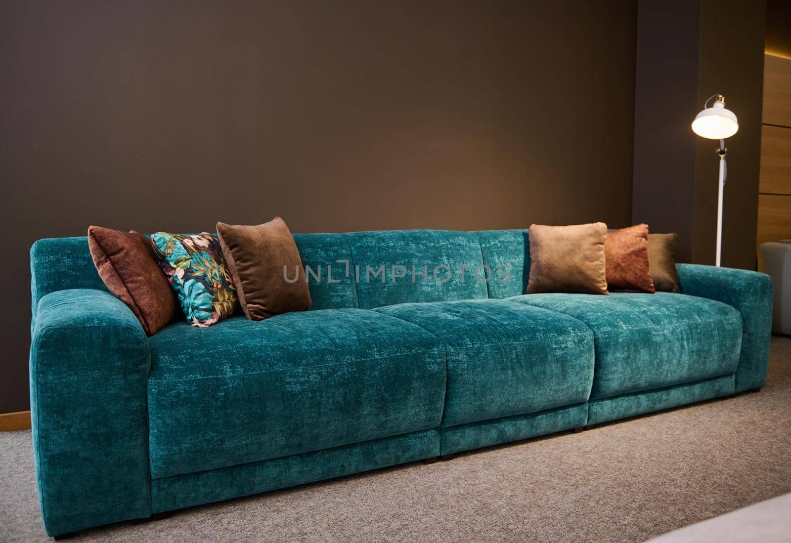 Exhibition of modern stylish upholstered furniture in the showroom of a furniture store. Focus on a turquoise soft velour sofa and brown pillows lit by a lamp against a brown wall background by artgf