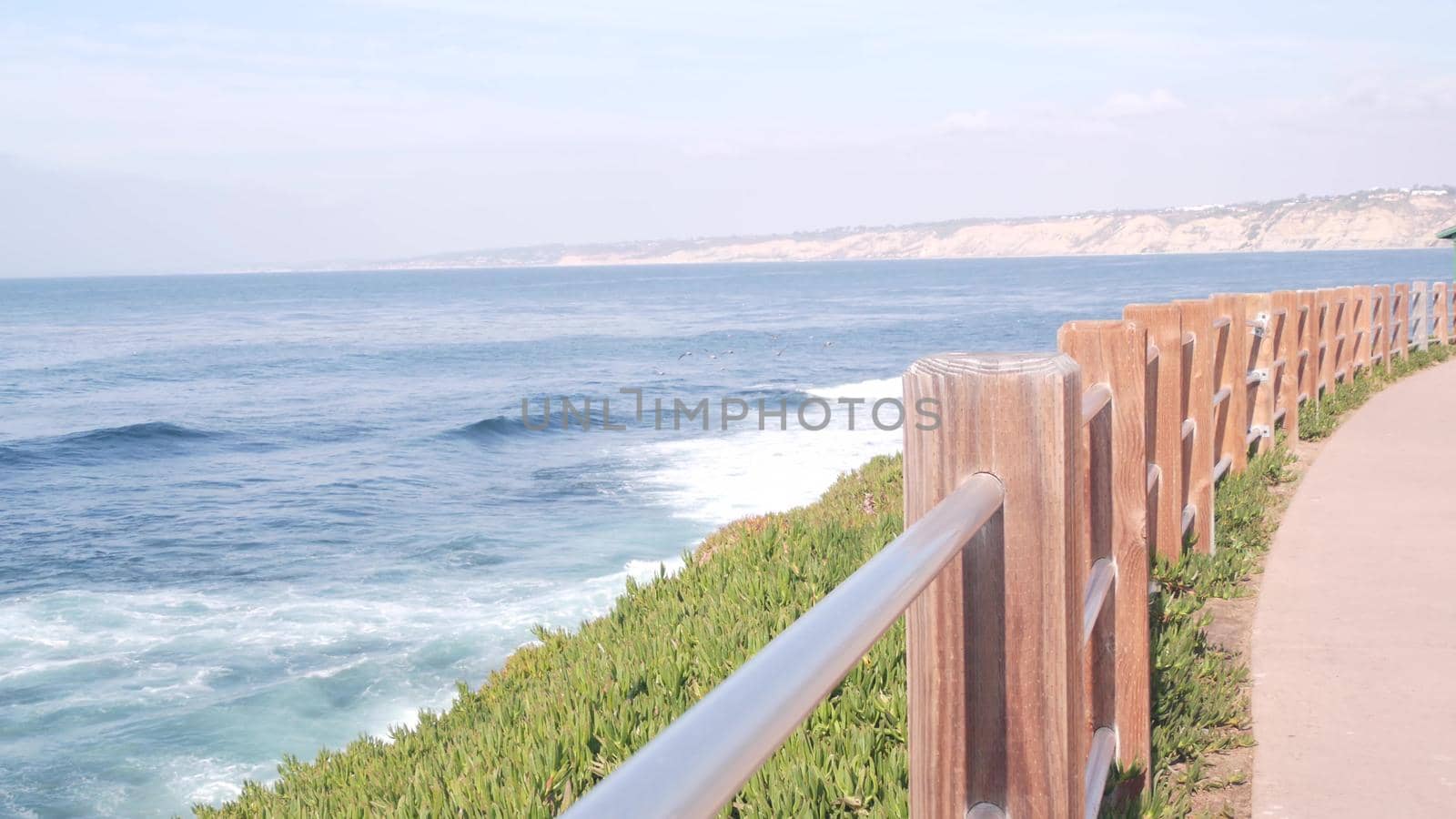Ocean waves crashing on beach, sea water surface from above, cliff or bluff, La Jolla shore waterfront promenade, California USA. Succulent green ice plant, pacific coast. Seascape view and railings.