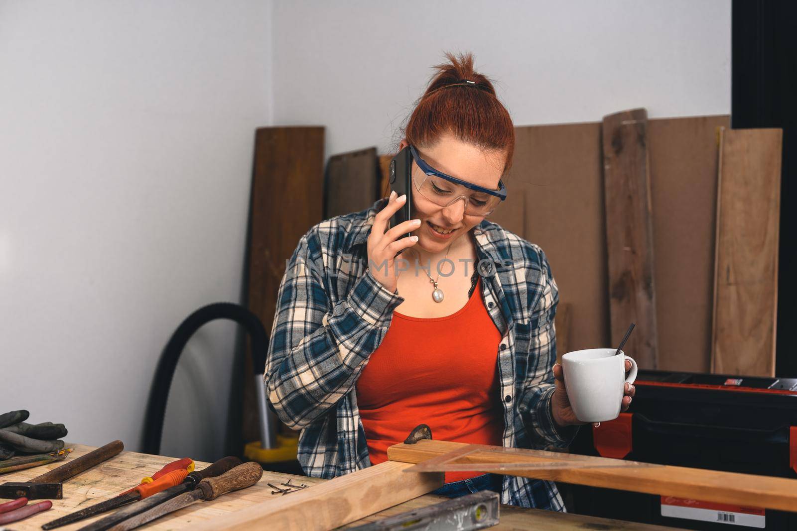 Young red-haired woman carpenter, concentrated and smiling, working on the layout of wood in a small carpentry shop, dressed in a blue checked shirt and a red t-shirt. Woman carpenter talking to a customer on her mobile phone, drinking a cup of coffee in the morning. Warm light indoors, background with wooden slats. Horizontal.