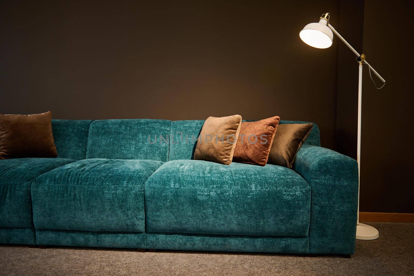 Exhibition of modern stylish upholstered furniture in the showroom of a furniture store. Focus on a turquoise soft velour sofa and brown pillows lit by a lamp against a brown wall background by artgf