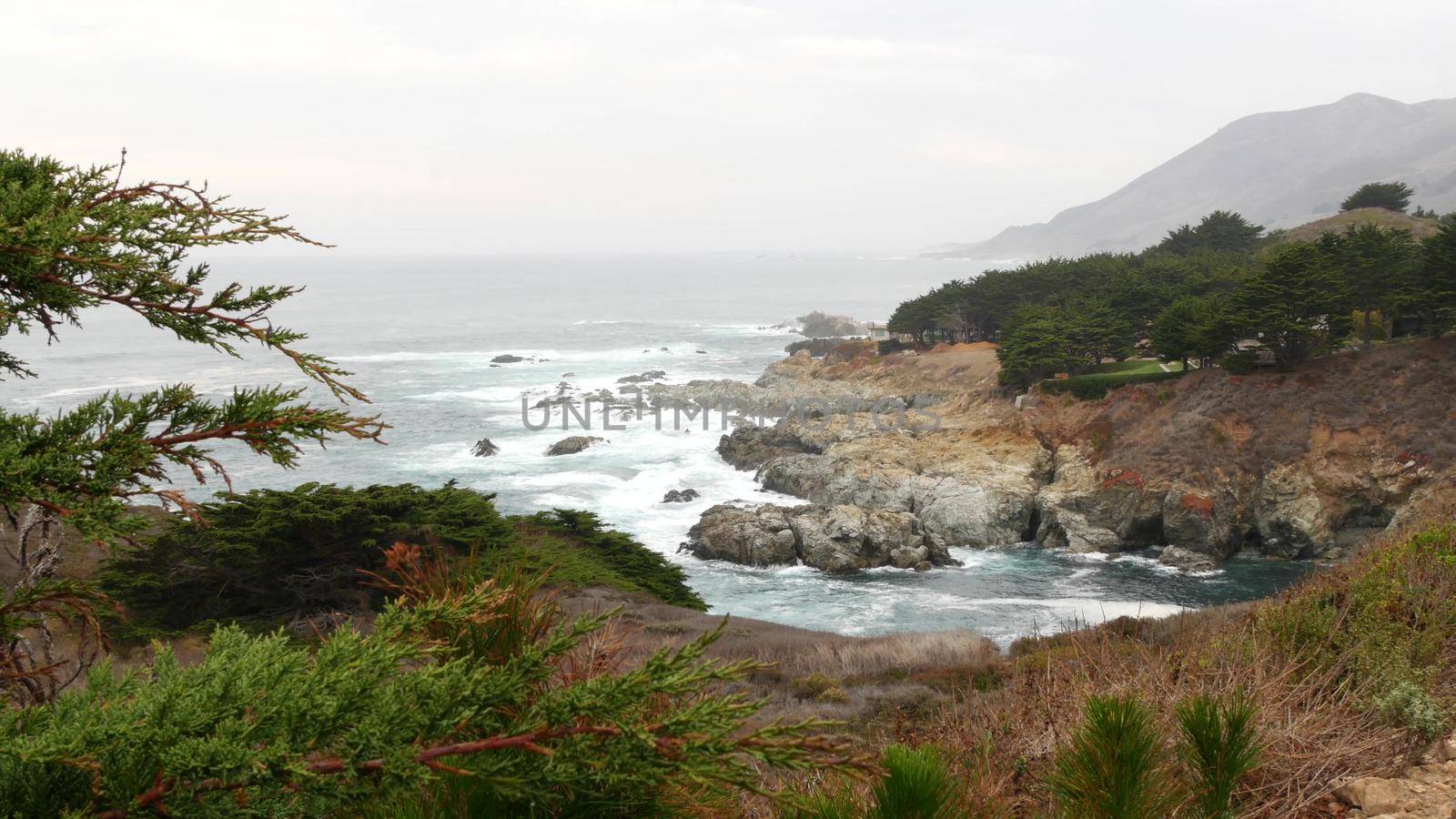Rocky craggy ocean shore, foggy misty weather. Sea water waves crashing on beach. California scenic landscape, Big Sur nature, USA. Pacific coast highway seascape viewpoint from cliff or steep bluff.