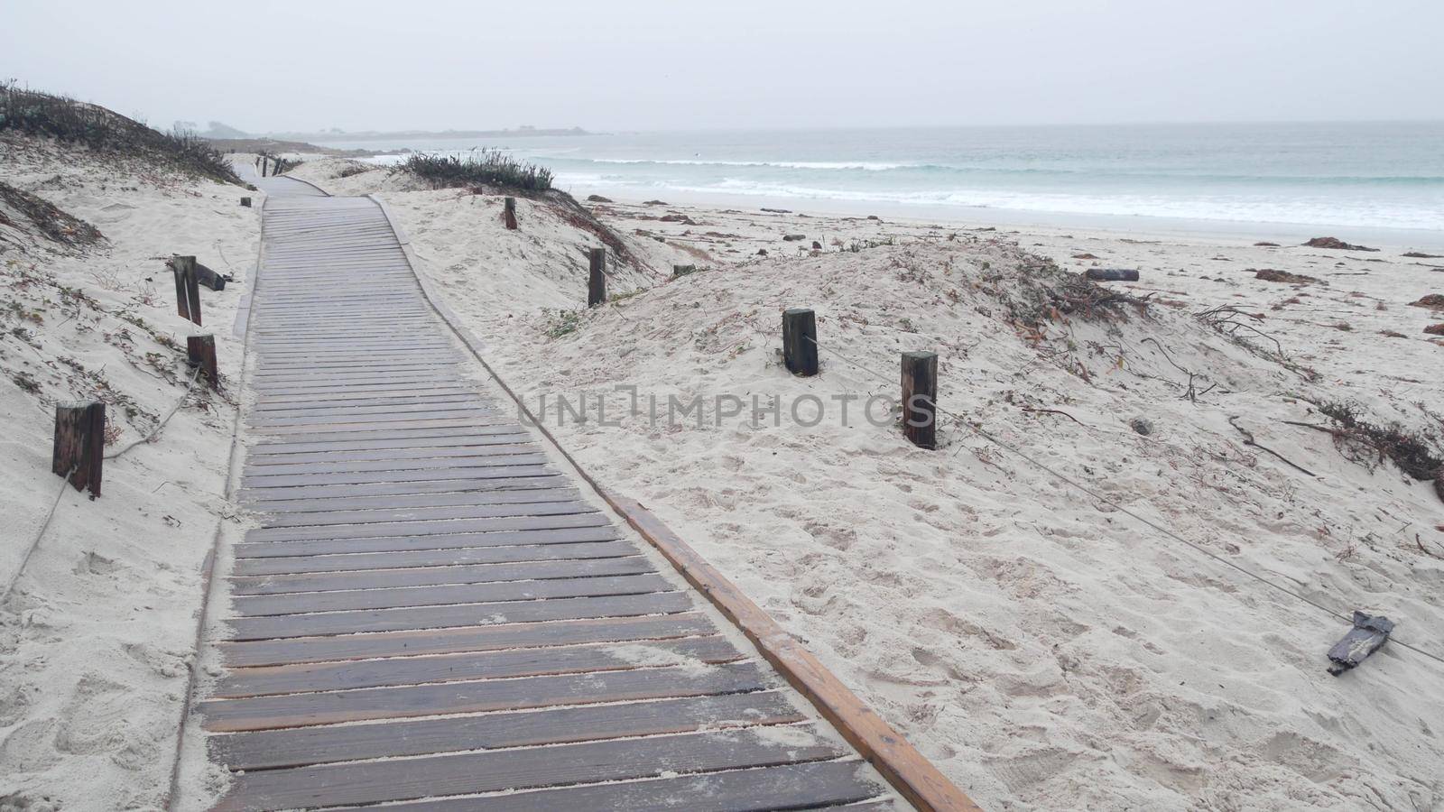 Ocean beach sandy dunes, Monterey nature, California misty coast, USA. Foggy rainy calm autumn or winter weather, grey cloudy sky. Trail path on shore near cold sea waves. Boardwalk from wooden planks