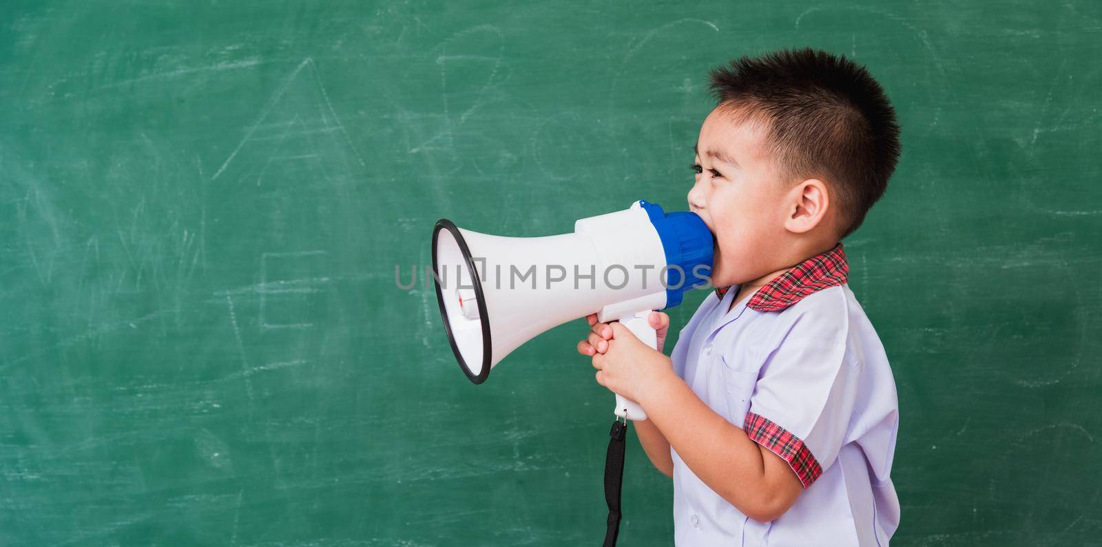 Back to School. Asian funny cute little child boy kindergarten preschool in student uniform speaking through megaphone against on green school blackboard, First time to school education concept