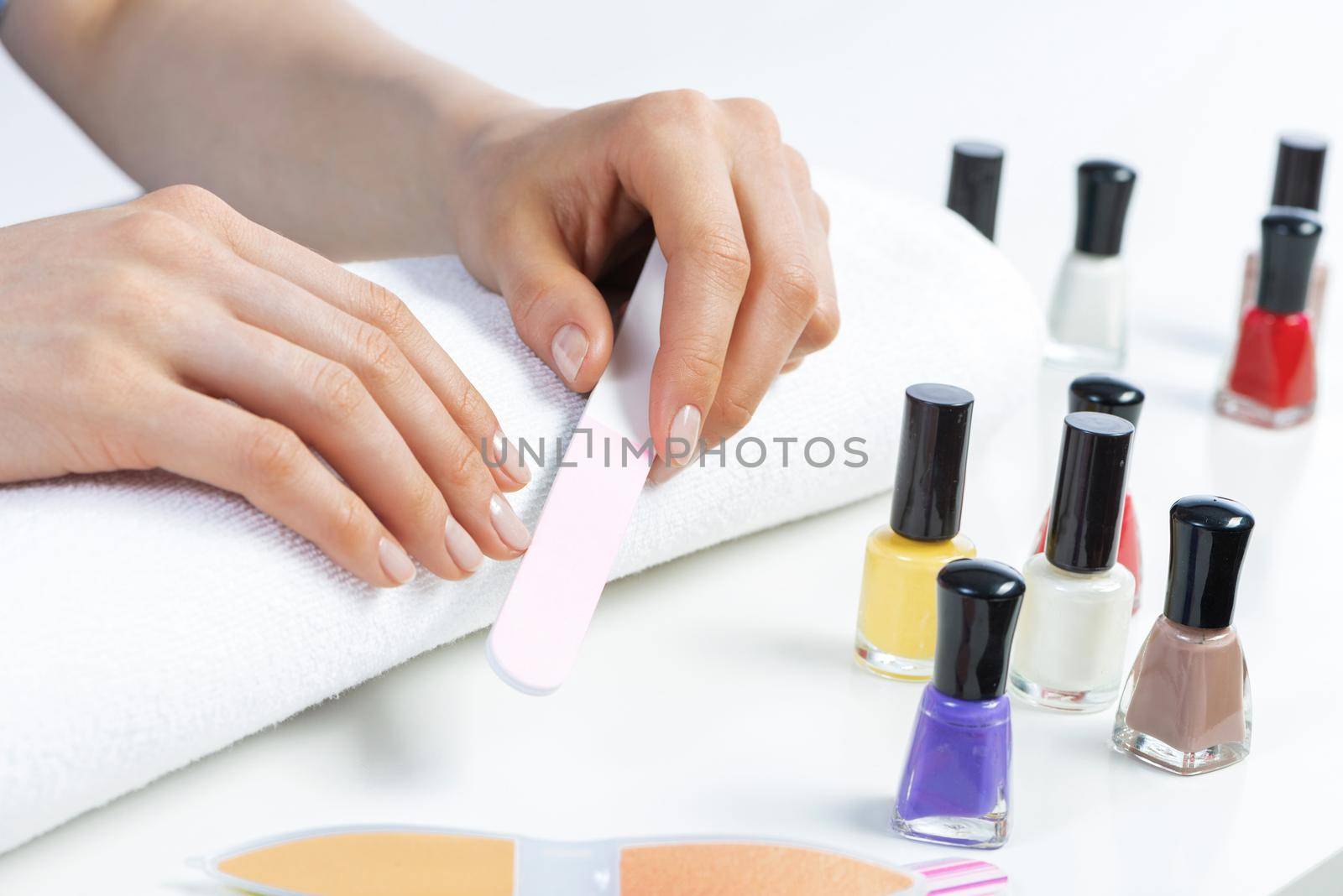 Woman using nail file and create perfect nails shape. Colorful nail polish bottles on table. Grinding female nails with nail file. Woman doing herself nail care procedure at home. Beauty and hygiene
