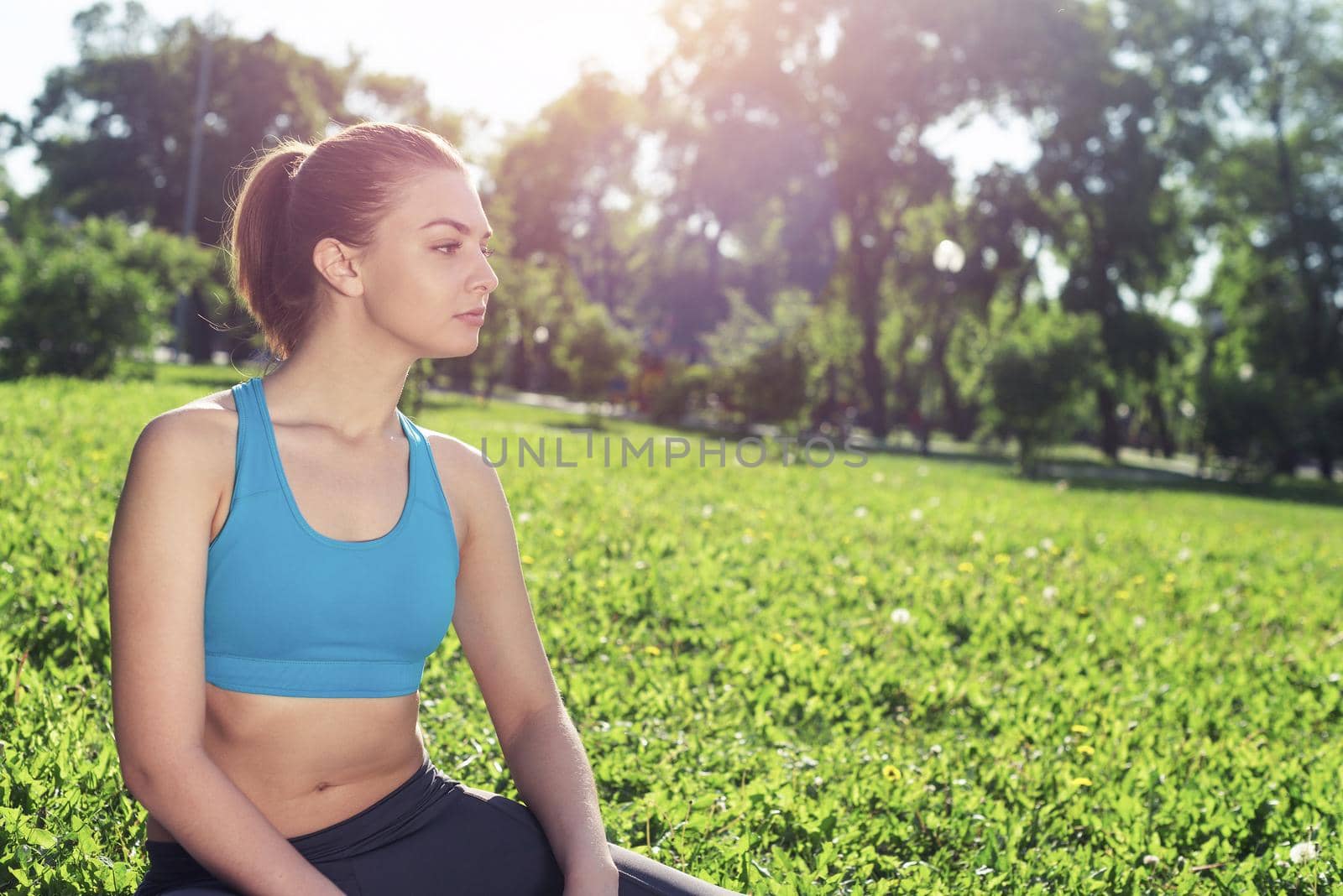 Beautiful smiling girl in activewear relax in park at sunrise. Portrait of young charming woman sitting on green grass. Training and meditation outdoor. Morning exercises and healthy lifestyle.