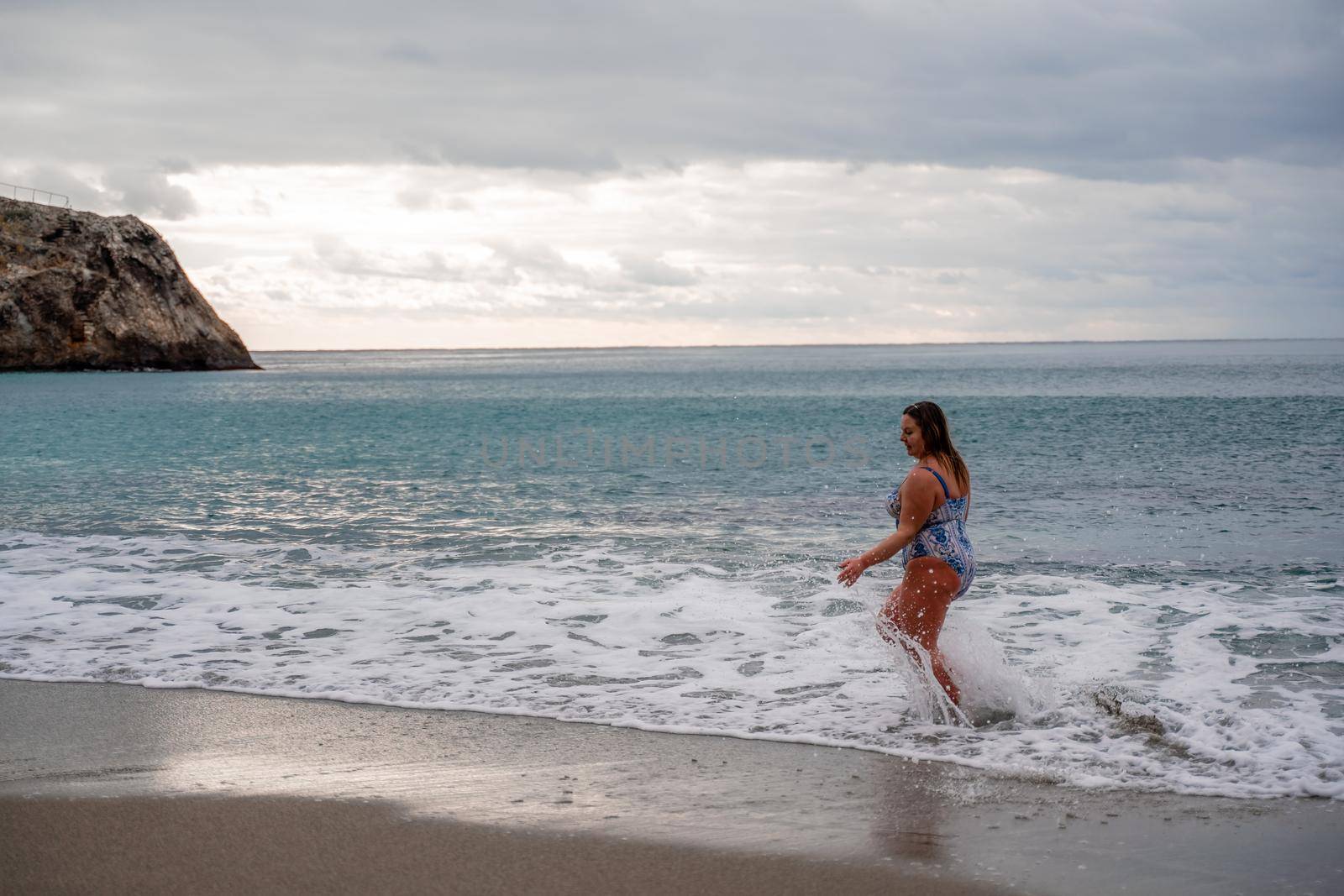 A plump woman in a bathing suit enters the water during the surf. Alone on the beach, Gray sky in the clouds, swimming in winter. by Matiunina