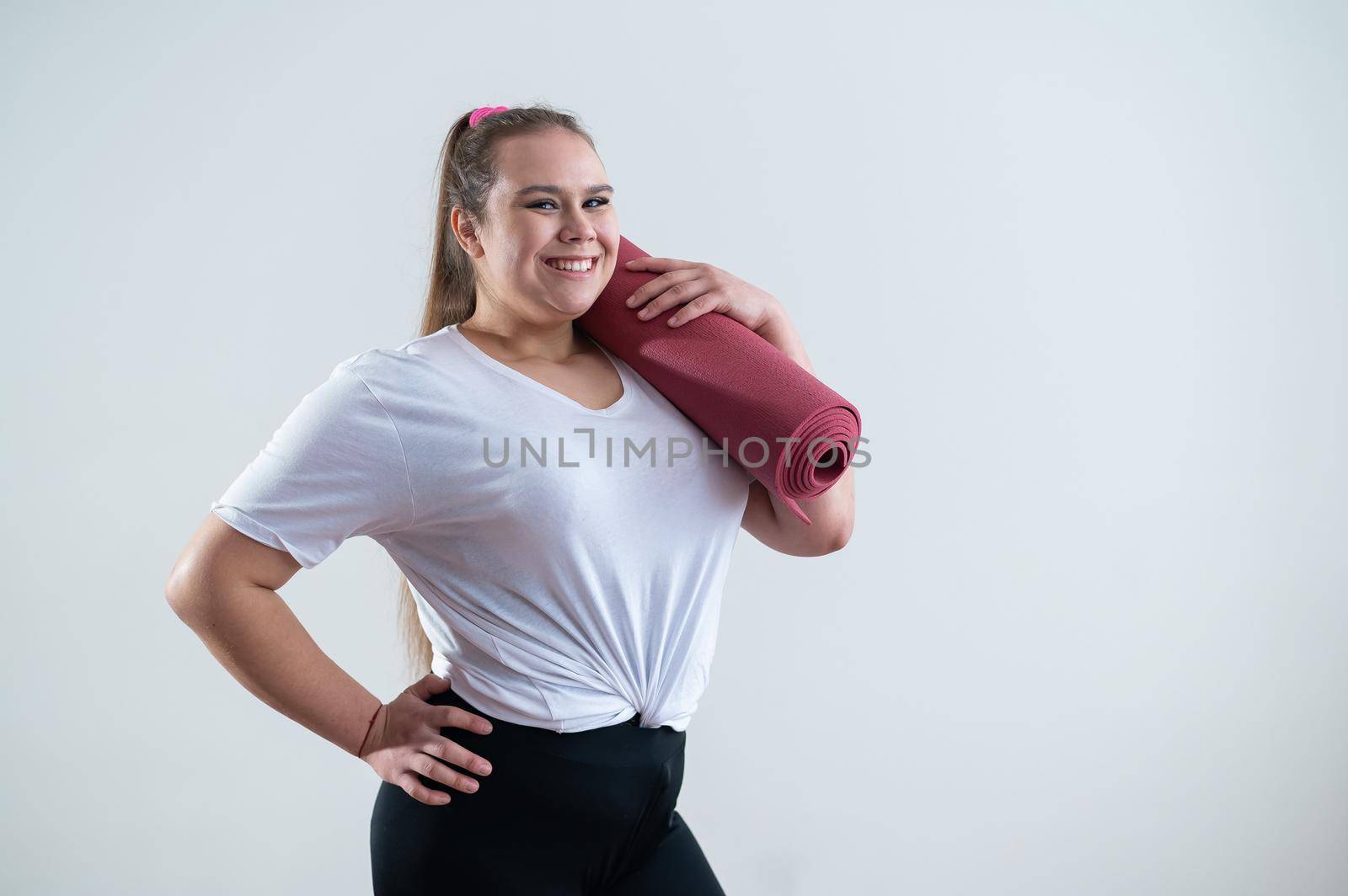 Young fat caucasian woman holding a sport mat. Charming plus size model in sportswear stands on a white background by mrwed54