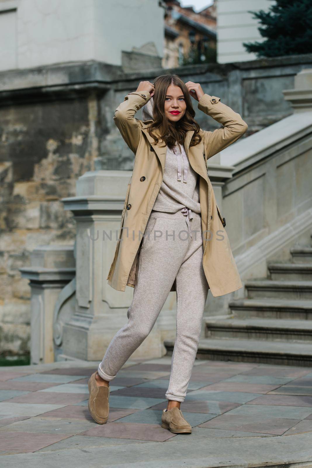 Portrait of fashionable women in beige sports suit, trench coat and stylish suede loafer posing on the stone stairs. street look fashion