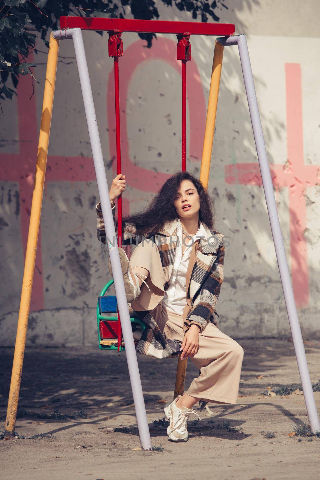 Closeup portrait of young beautiful fashionable woman wearing checkered long coat, beige pants and white blouse . Lady posing on city building background. Female fashion.