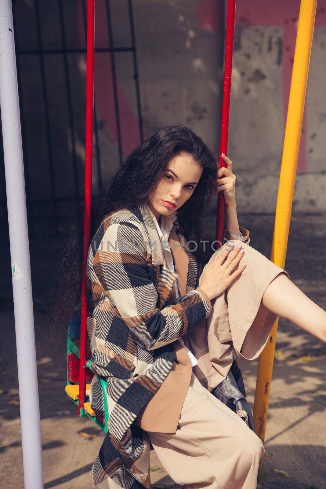 Closeup portrait of young beautiful fashionable woman wearing checkered long coat, beige pants and white blouse . Lady posing on city building background. Female fashion.