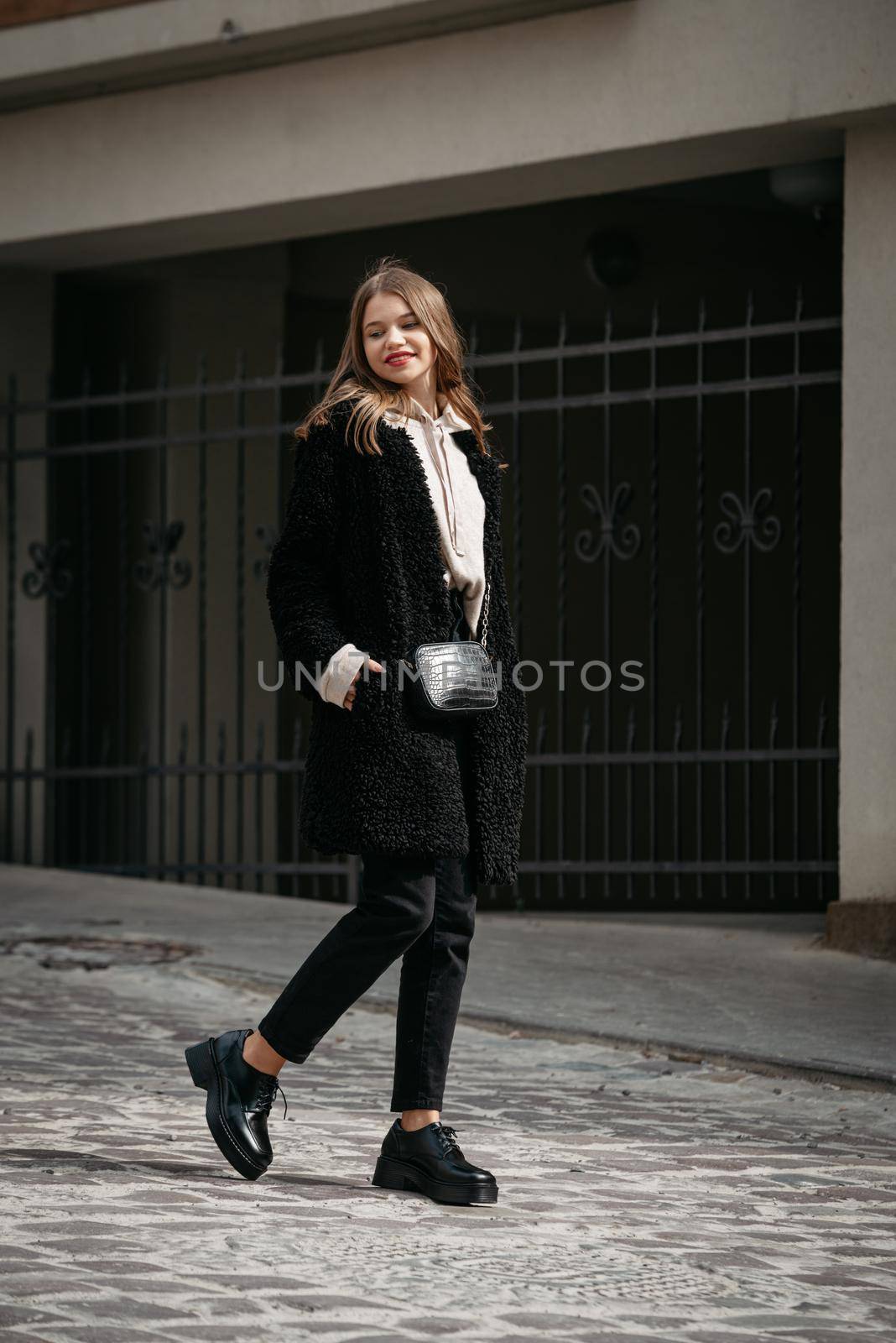 Fashion photo of young woman in black fur coat, jeanse and shoes at city street. beige jacket with a hood