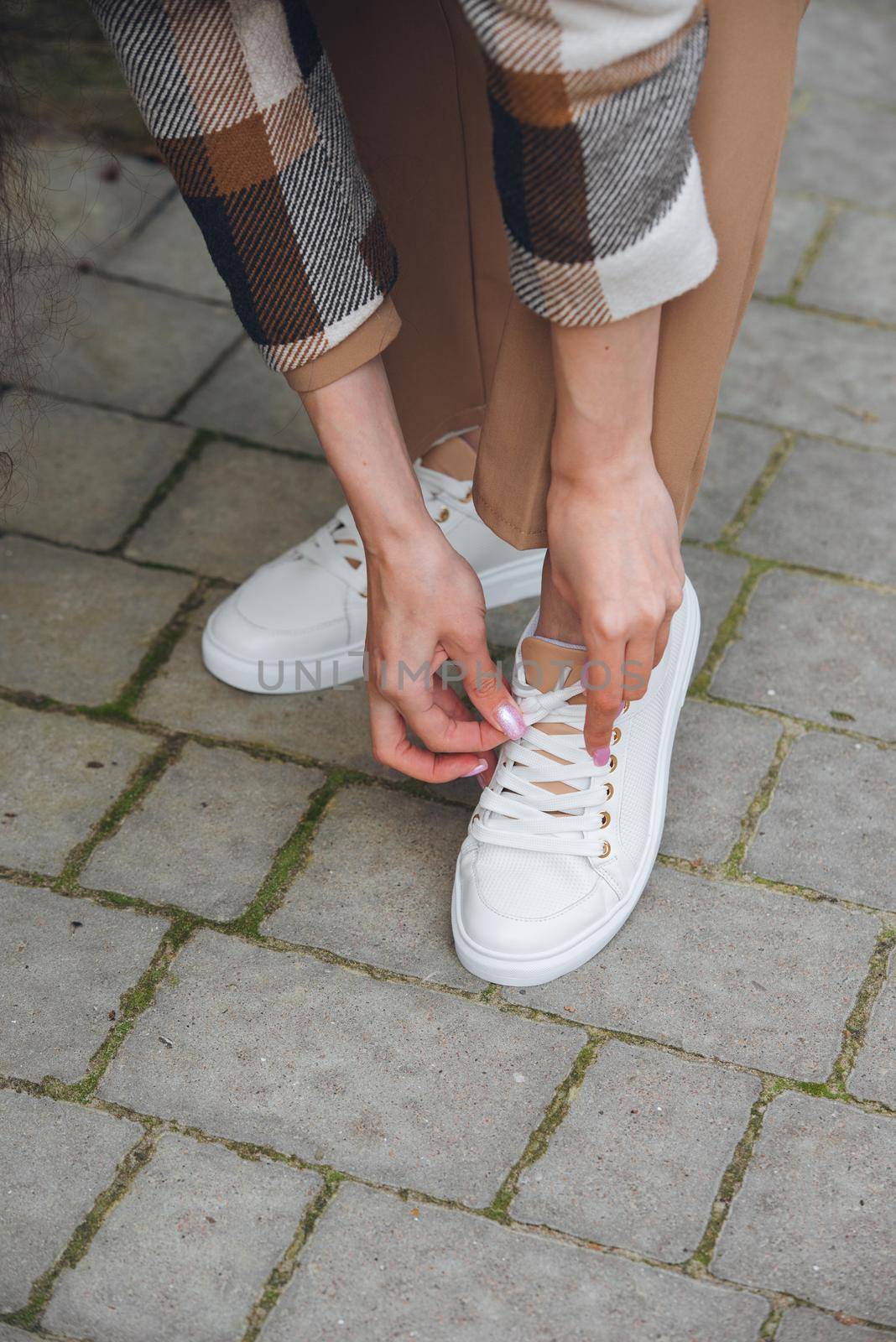 Closeup photo of young woman wearing checkered long coat, and beige pants . Lady posing on city street. Women hands ties laces on sneakers