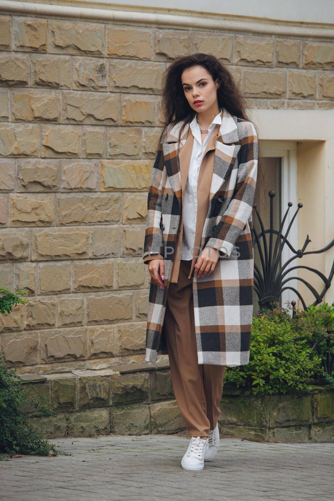 Closeup portrait of young beautiful fashionable woman wearing checkered long coat, beige pants and white blouse . Lady posing on city street.. Female fashion.