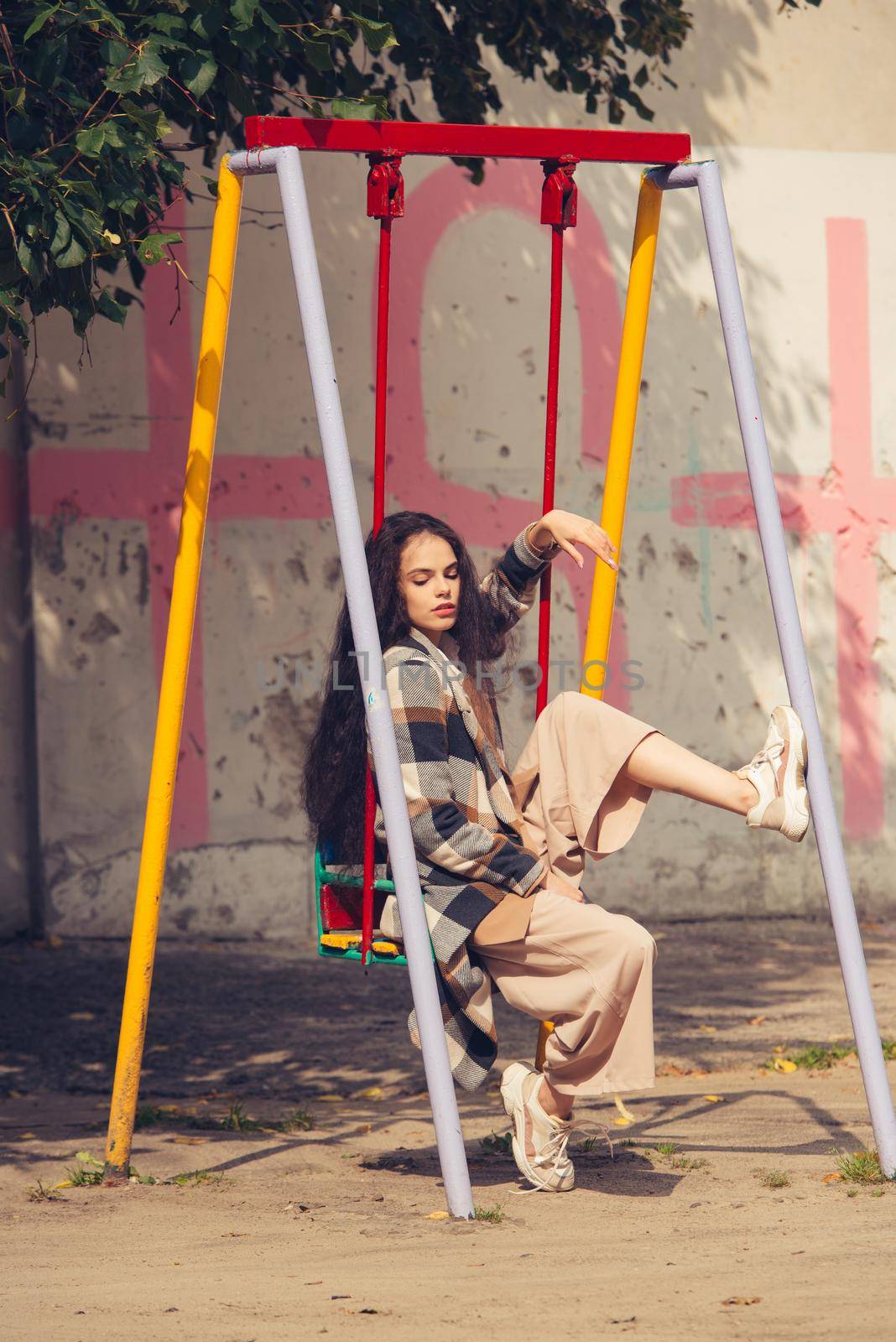 Closeup portrait of young beautiful fashionable woman wearing checkered long coat, beige pants and white blouse . Lady posing on a swing by Ashtray25