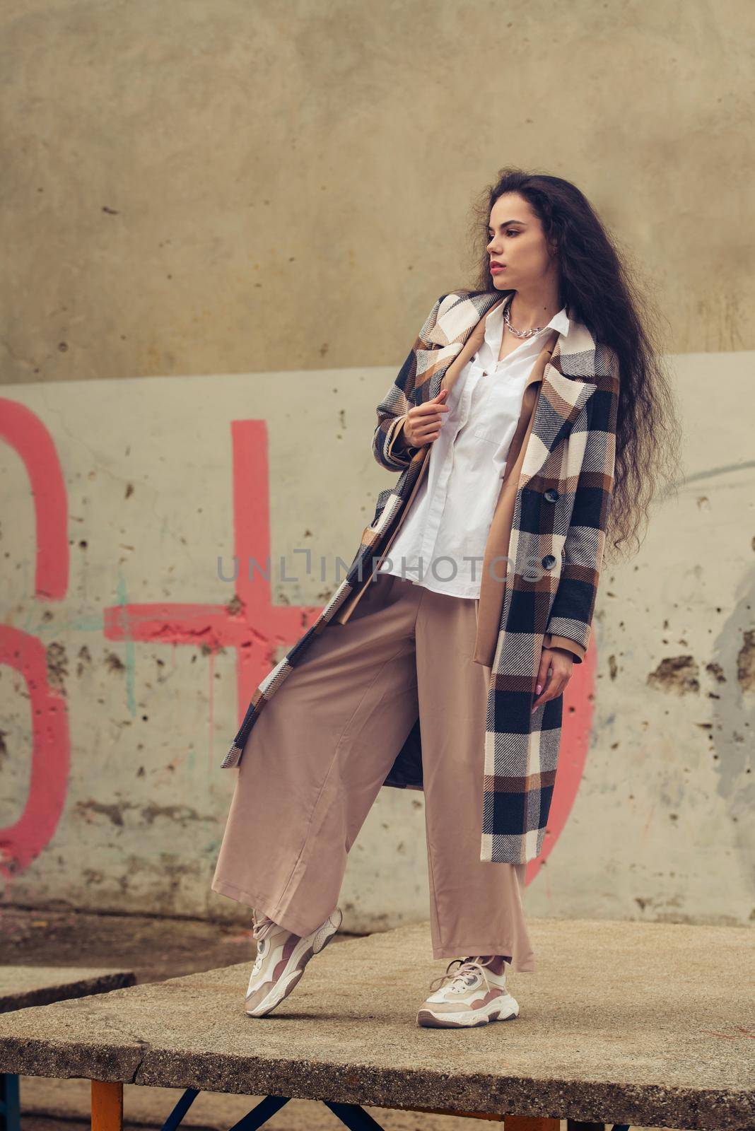 Closeup portrait of young beautiful fashionable woman wearing checkered long coat, beige pants and white blouse . Lady posing on city building background. Female fashion.