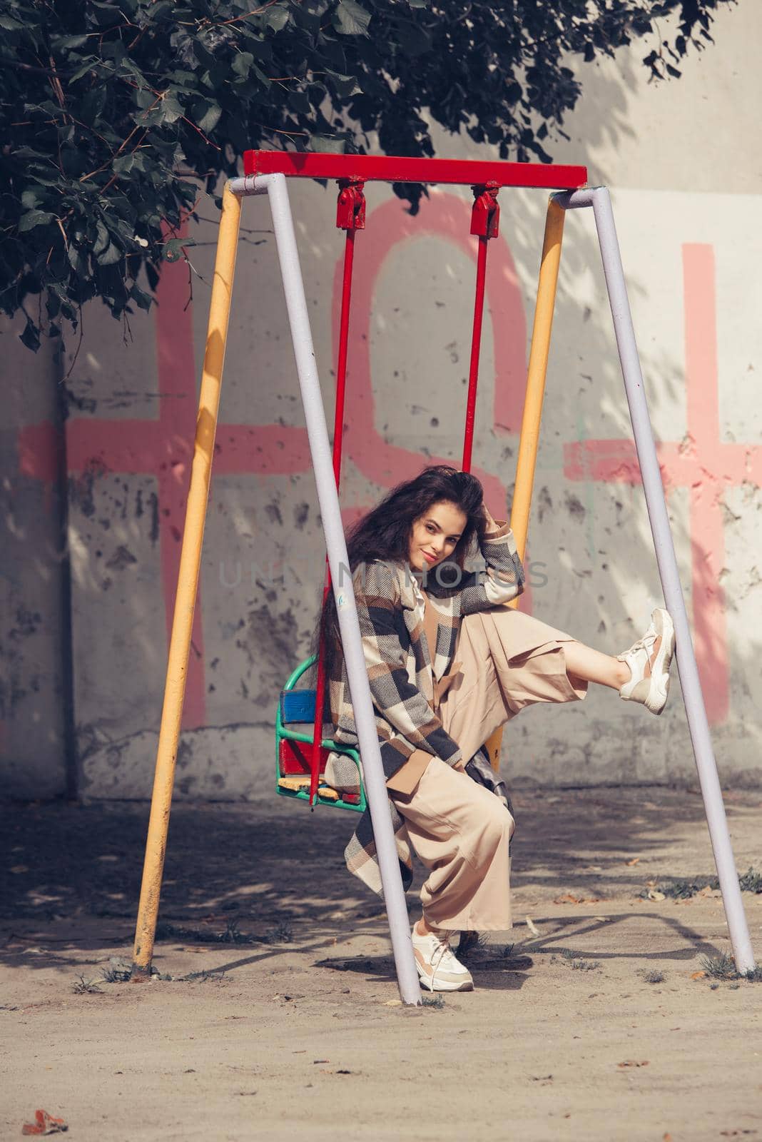 Closeup portrait of young beautiful fashionable woman wearing checkered long coat, beige pants and white blouse . Lady posing on city building background. Female fashion.