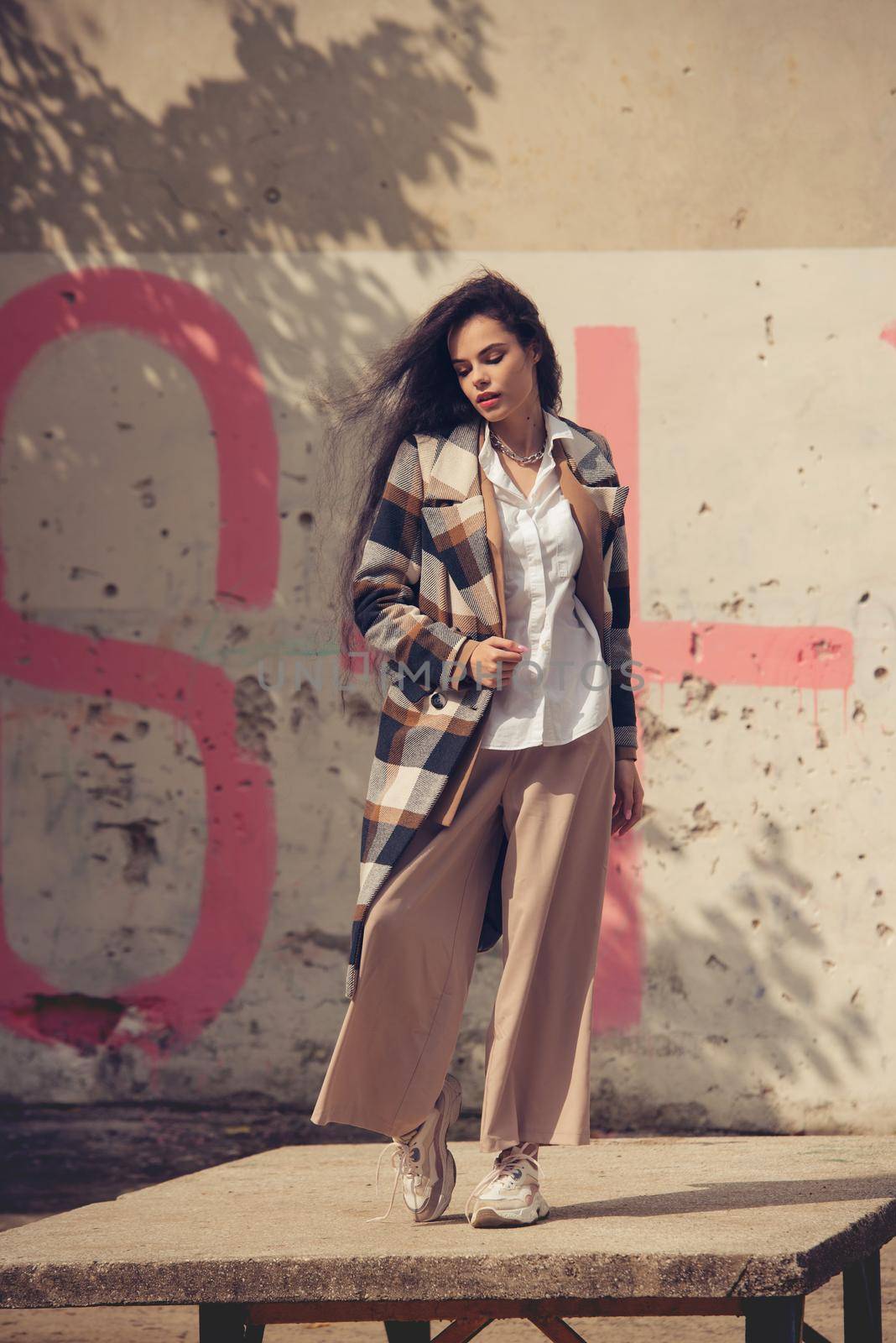 Closeup portrait of young beautiful fashionable woman wearing checkered long coat, beige pants and white blouse . Lady posing on city building background. Female fashion.