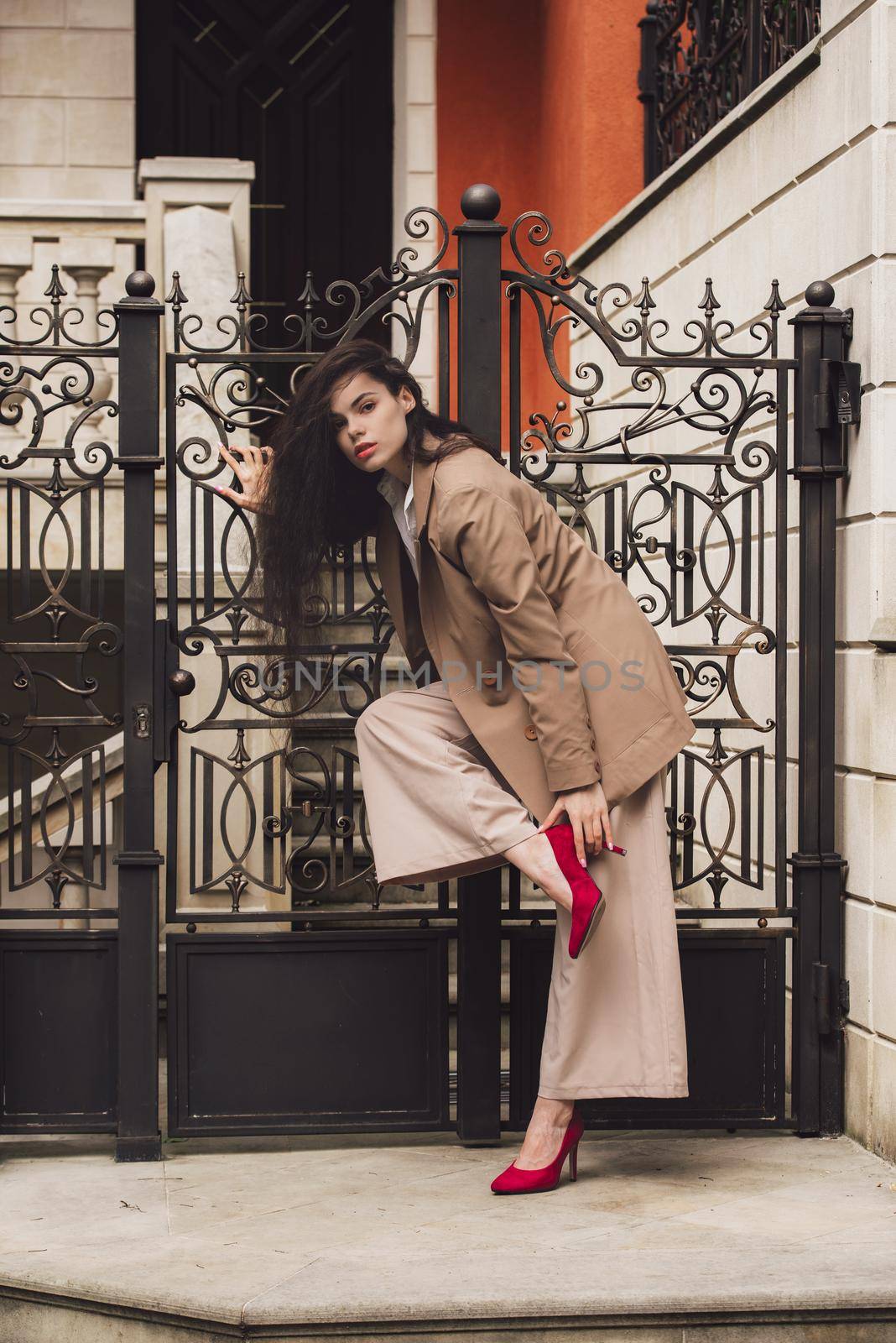 Close up portrait of young beautiful woman with long brunette curly hair posing against building background. woman wearing a white blouse and beige pants and a jacket