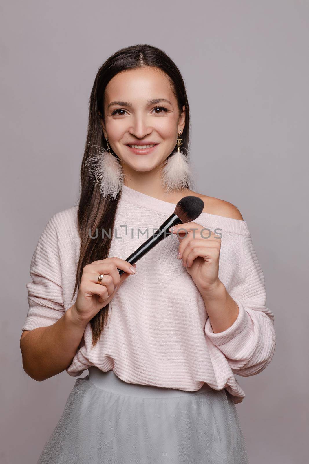 Beautiful cute girl in sweater with one naked shoulder posing at camera. Young make up artist holding one hand on belly and in other big brush. Happy brunette lady with pink feather earrings smiling.