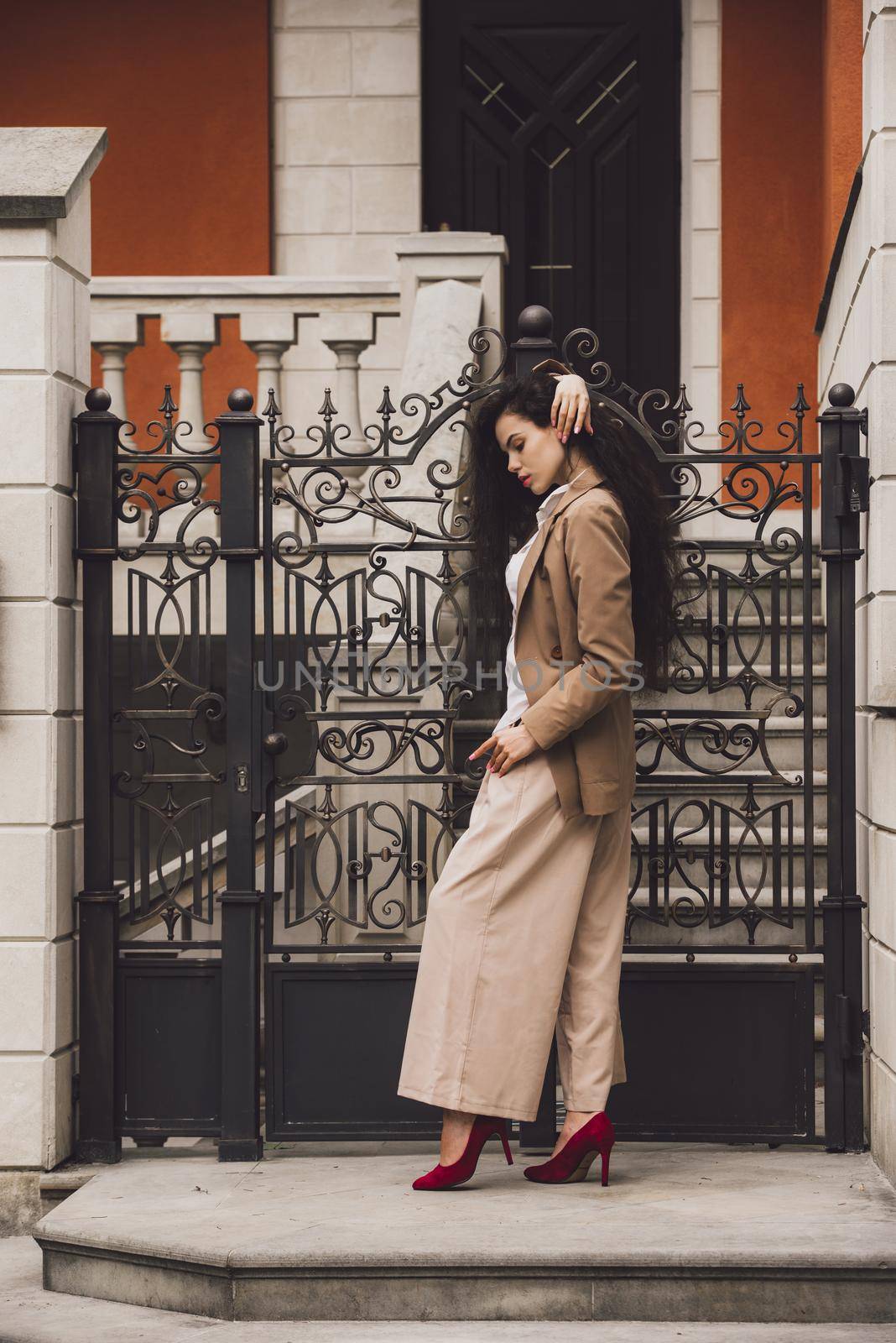 Close up portrait of young beautiful woman with long brunette curly hair posing against building background. woman wearing a white blouse and beige pants and a jacket