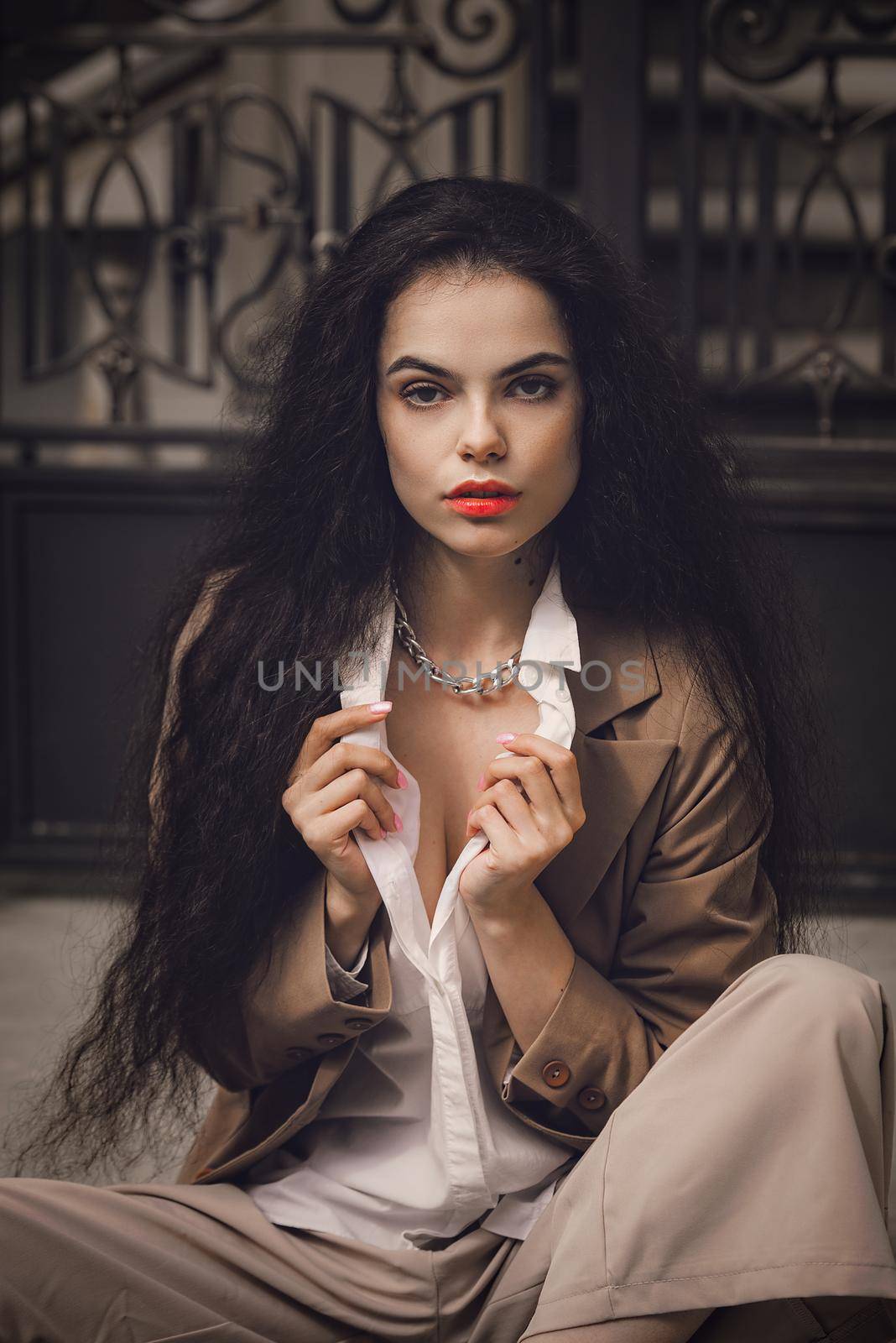 Close up portrait of young beautiful woman with long brunette curly hair posing against building background. woman wearing a white blouse and beige pants and a jacket