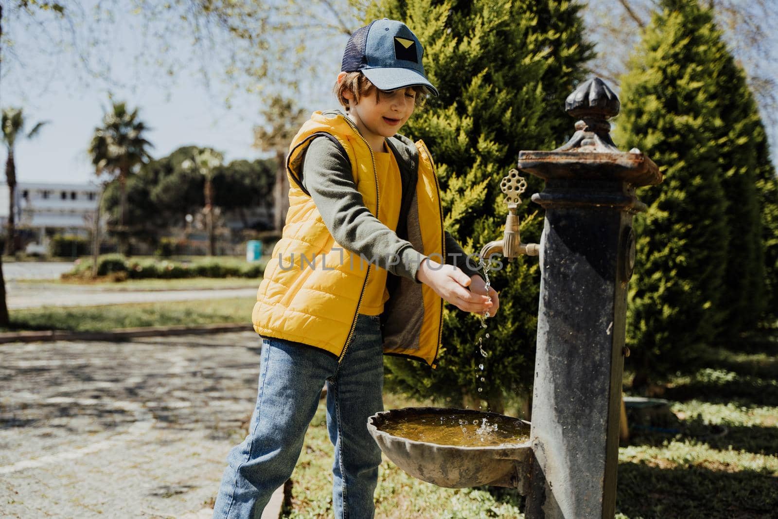 Kid schoolboy in casual clothing washing his hands in the street old fashioned drinking water fountain. Boy in blue jeans and yellow vest playing with water from drinking sprinkler in city park.