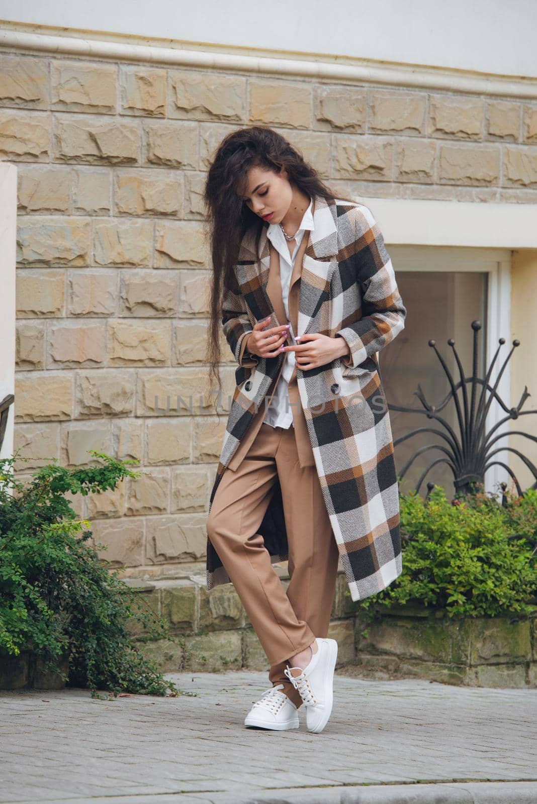 Closeup portrait of young beautiful fashionable woman wearing checkered long coat, beige pants and white blouse . Lady posing on city street.. Female fashion.