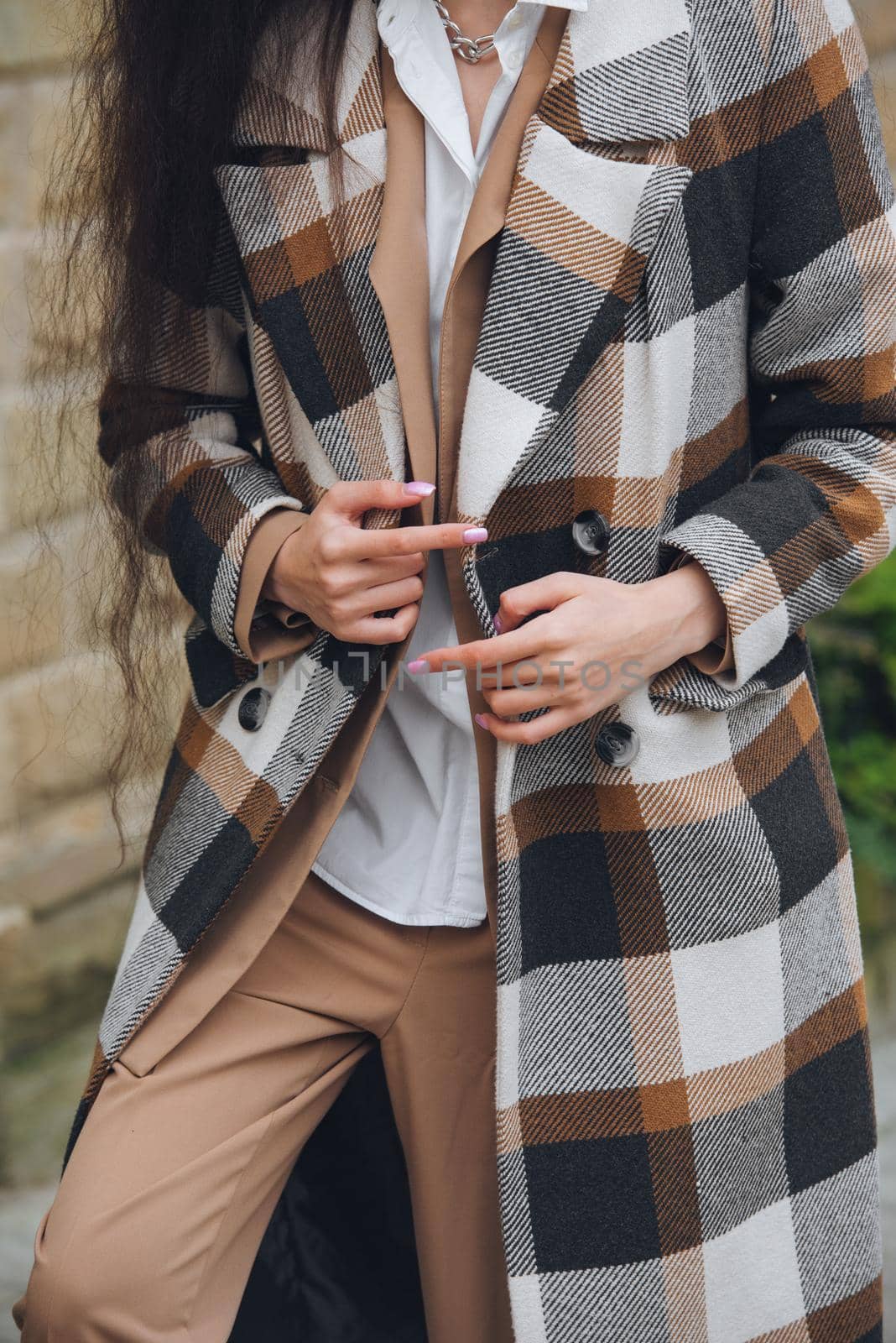 Closeup portrait of young beautiful fashionable woman wearing checkered long coat, beige pants and white blouse . Lady posing on city street.. Female fashion.