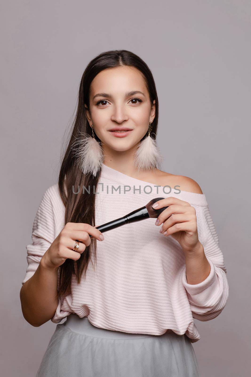 Beautiful cute girl in sweater with one naked shoulder posing at camera. Young make up artist holding one hand on belly and in other big brush. Happy brunette lady with pink feather earrings smiling.