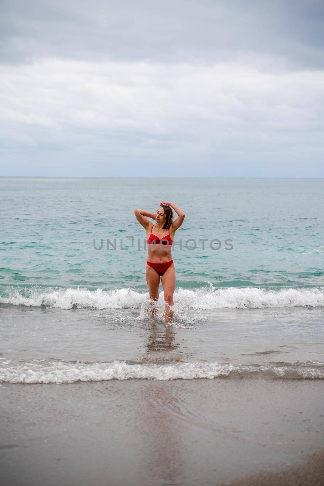 A middle-aged woman with a good figure in a red swimsuit on a pebble beach, running along the shore in the foam of the waves.