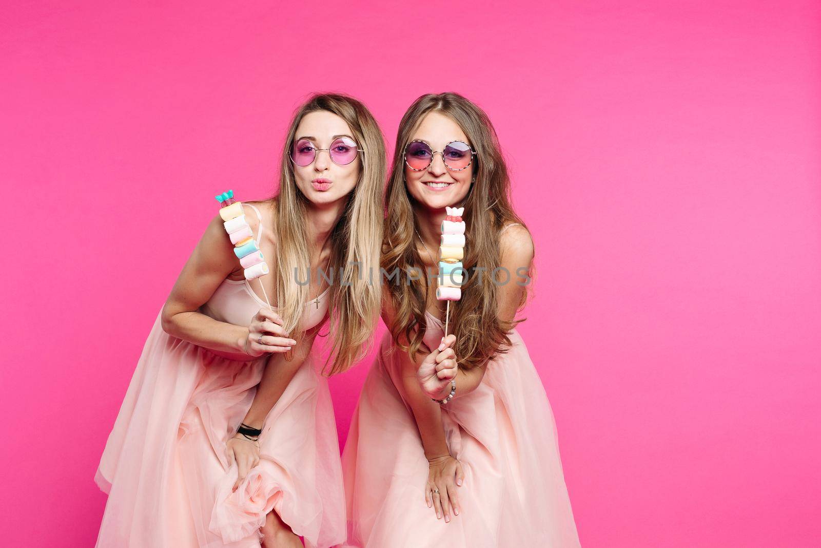 Studio portrait o two beautiful and sweet girls, in pink sunglasses and looks like princes or candies, sending kiss at camera. Pretty women showing marshmallows on stick and having fun. Fashion.
