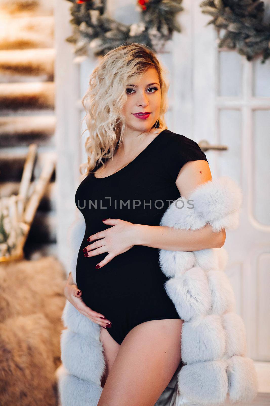 View of beautiful blonde pregancy mom lying, posing at studio, looking, smiling at camera. Mother expacting for child, posing at decorated studio for new year and Christmas holiday. Woman wearing in white for, with make up for x mas paty.