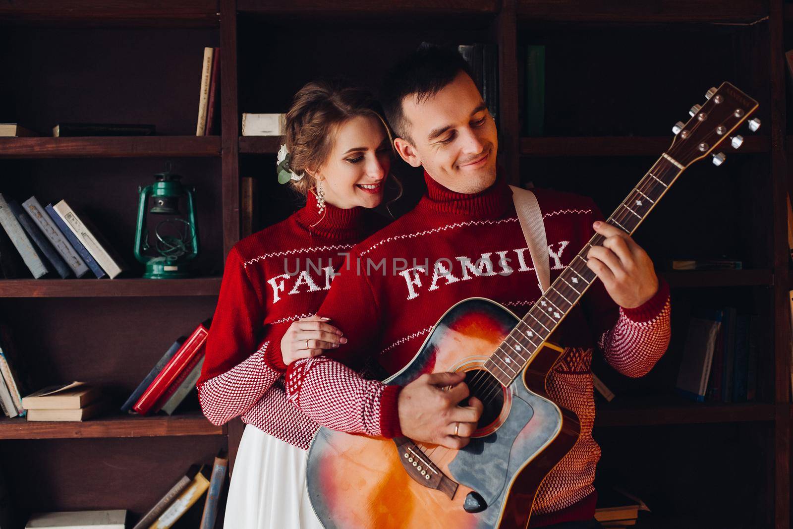 Beautiful smiling couple in red sweaters listening music, girlfriend leaning on handsome boyfriend, when he playing at gitar. Celebrating new year and xmas. Romantic winter holidays.